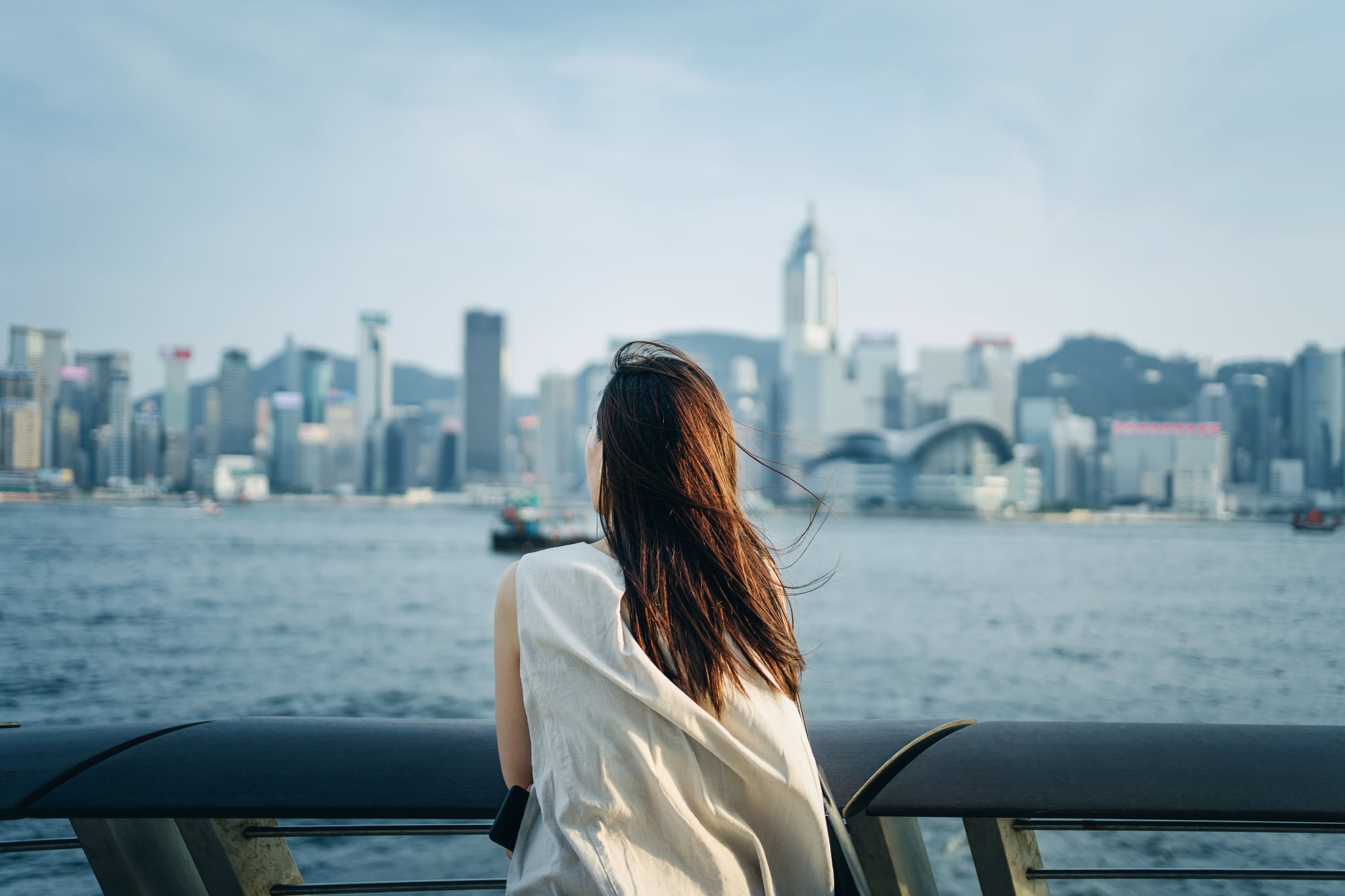 Relaxing young Asian woman traveller looking out over promenade of Victoria Harbour enjoying the gentle breeze and the beautiful cityscape at dusk
