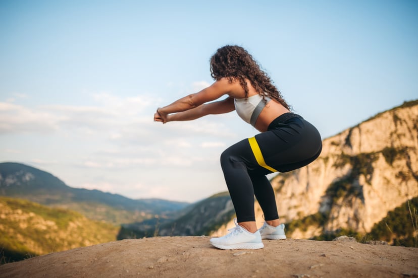 Young caucasian beautiful fit woman standing on top of a mountain and doing squats with resistance band.