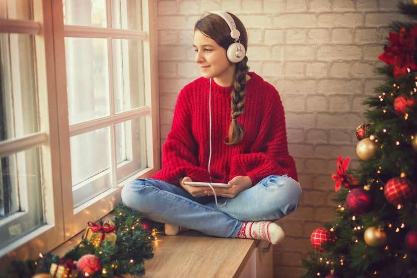 Teenage girl at home for holiday, she sitting by the window using digital tablet and listening to music in cozy Christmas atmosphere
