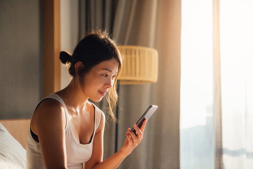 Portrait of young Asian woman holding smartphone while waking up in bed in the morning.