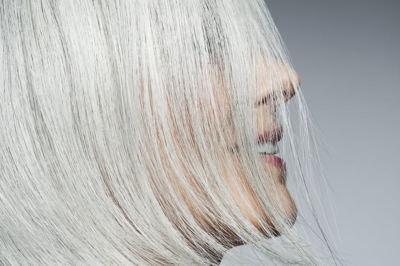 Woman with medium length, silvery, grey hair covering face in front of grey background in a profile position, cropped.
