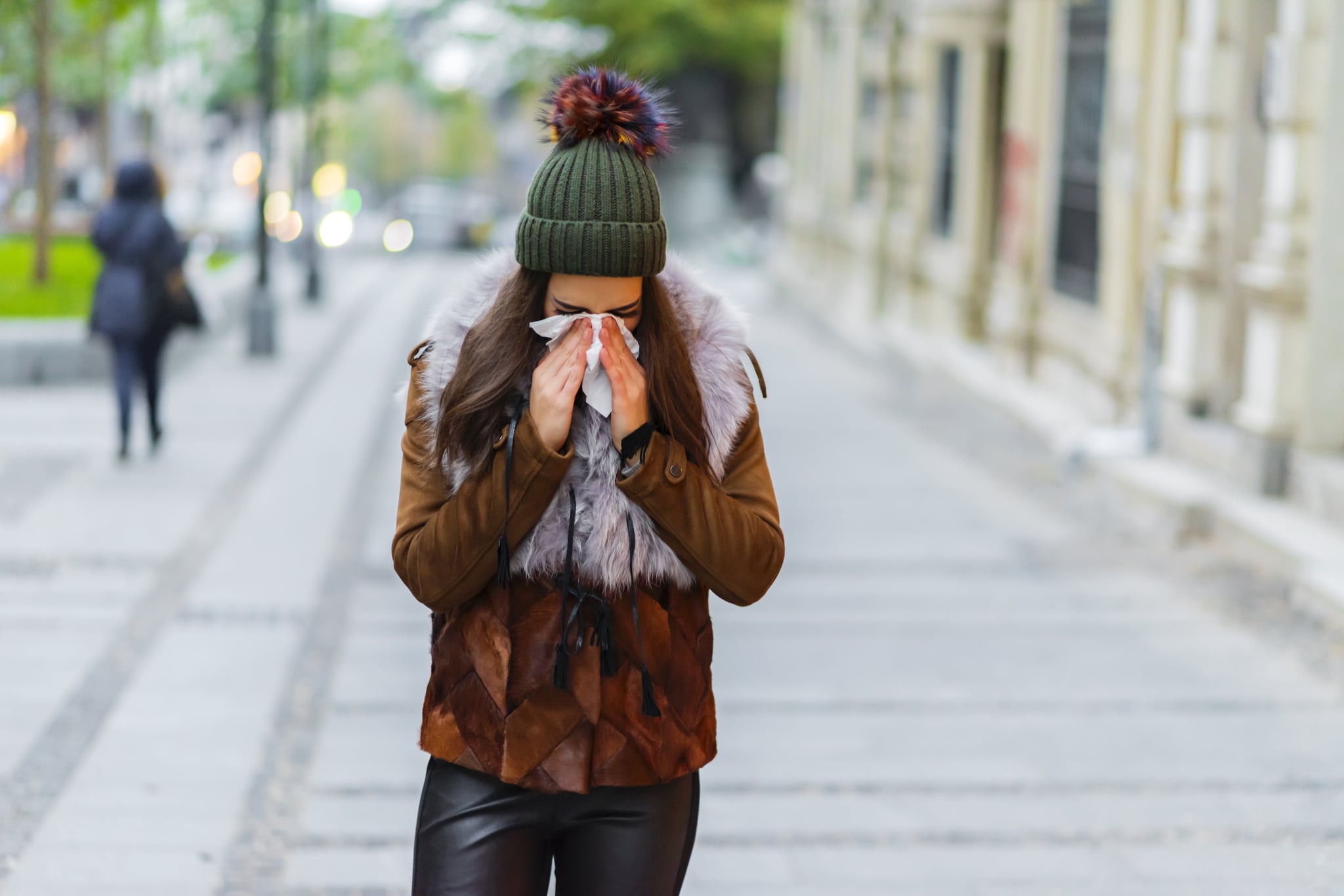 Photo of young Woman standing on the street blowing her nose. Healthcare, flu and people concept - Ill woman blowing nose to paper napkin.