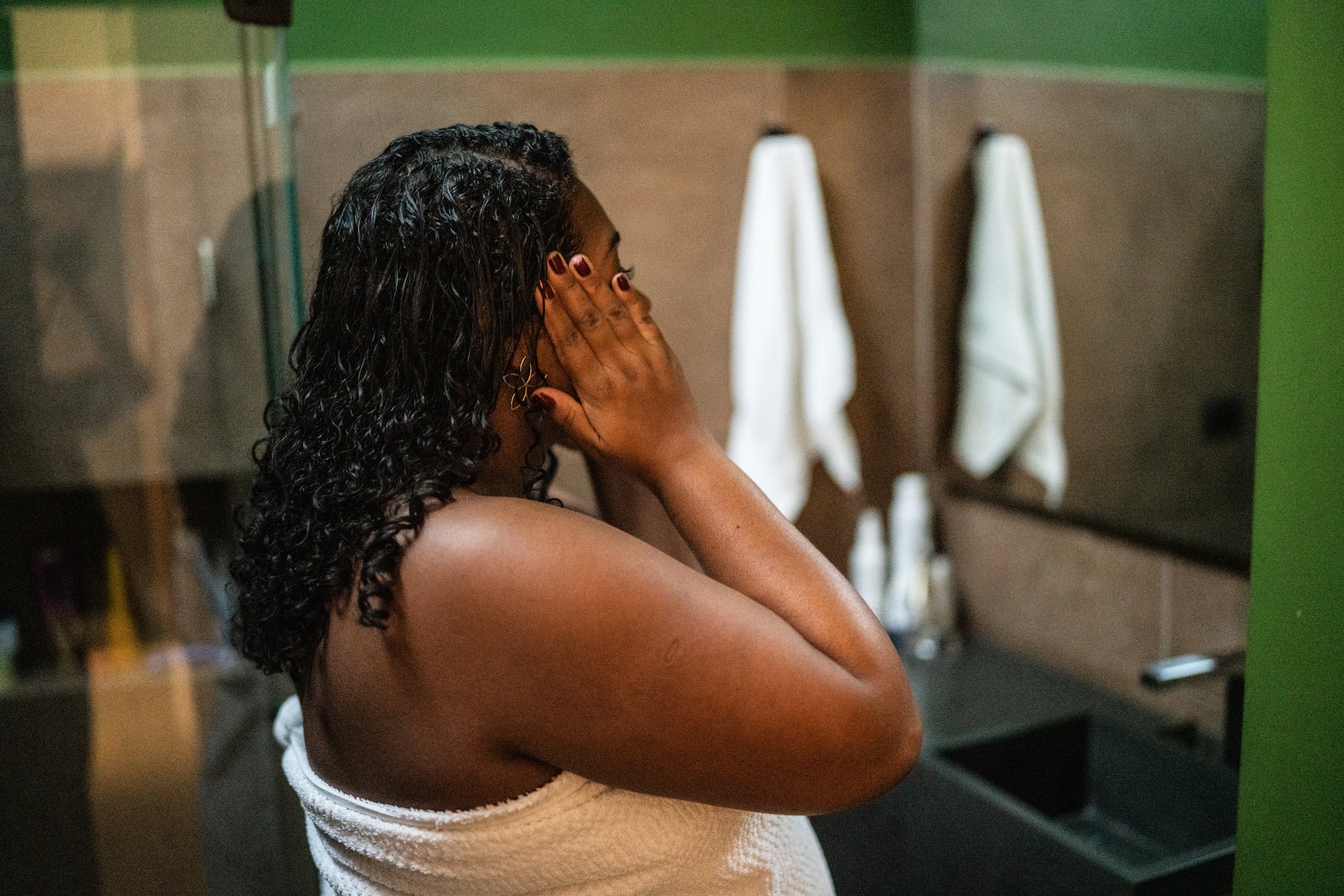A Black woman stands in her bathroom and looks into a mnirror as she applies face lotion.