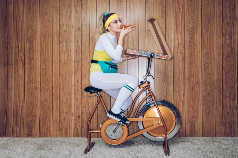A woman wearing exercise clothing styled after the 1980's and 1990's pedals hard on a stationary fitness bike in a vintage room, complete with shag carpet and wood paneling on the walls. She wears a leotard and a fanny pack and eats from a large box of pi