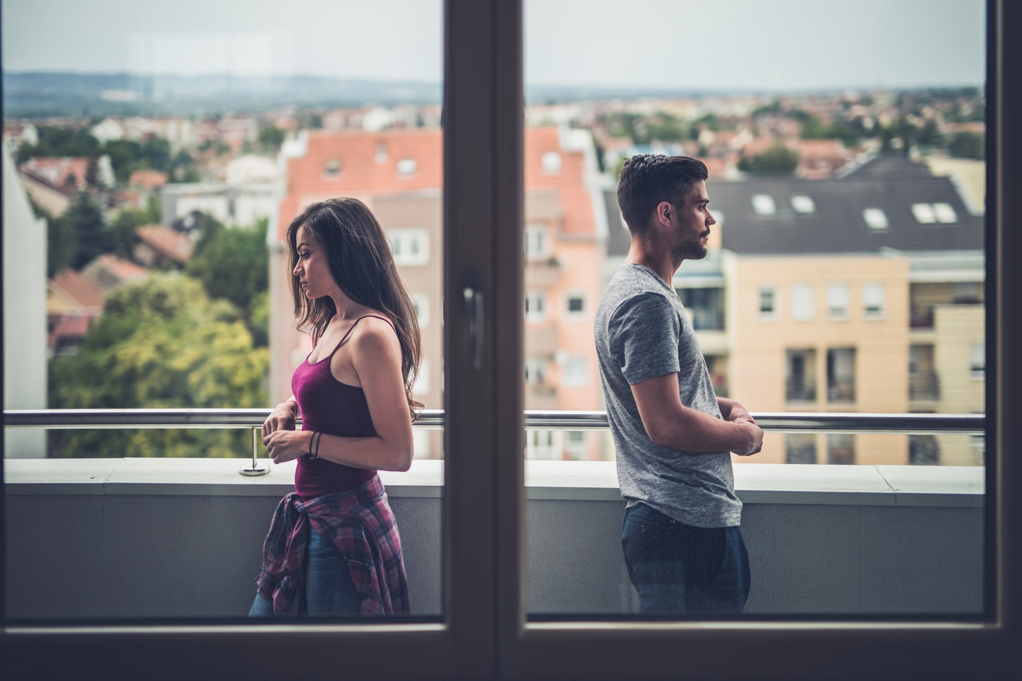 Sad couple standing back to back on a penthouse terrace while having problems in their relationship. The view is through window.
