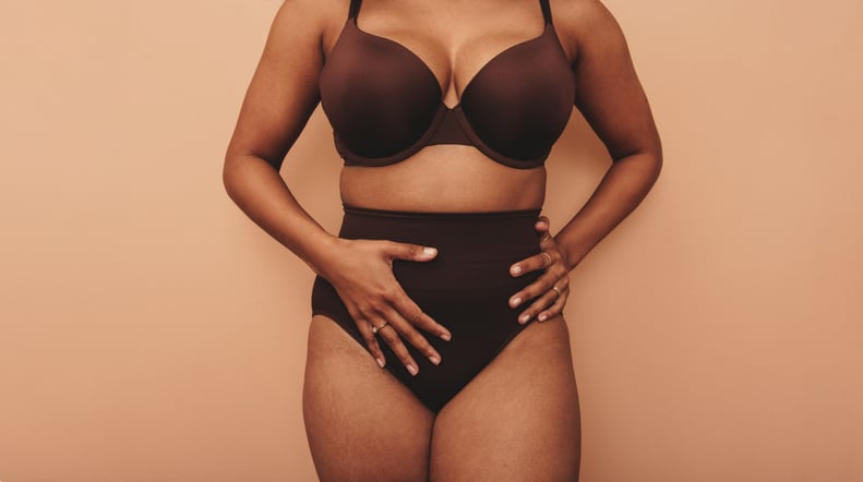 Unrecognizable young woman embracing her midsection in a studio. Anonymous young woman wearing brown underwear against a studio background. Self-confident young woman embracing her natural body.