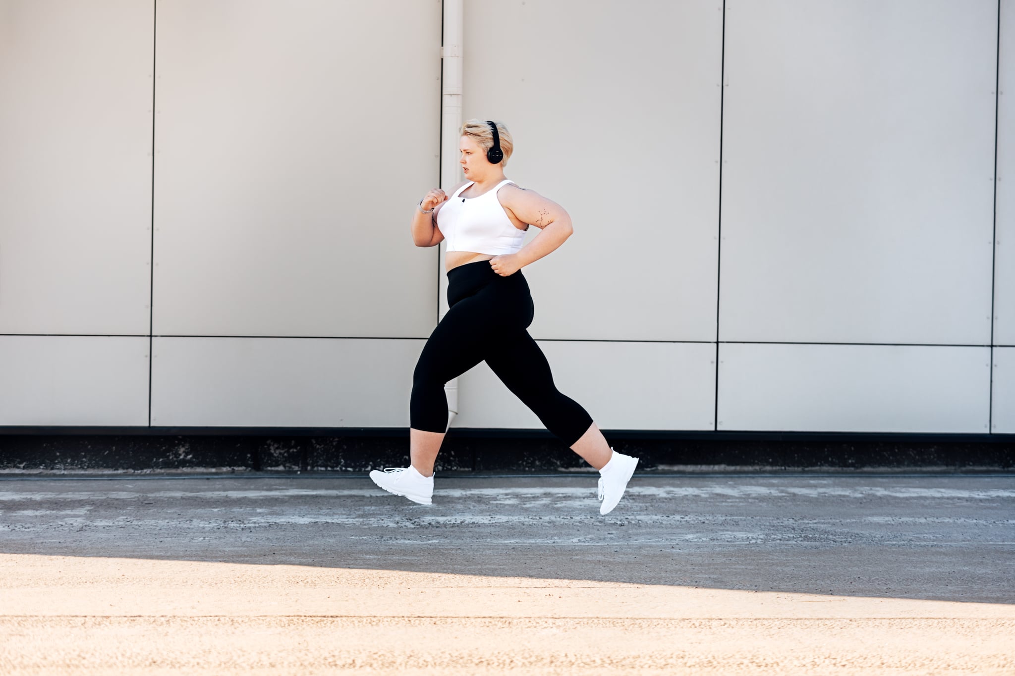 Side view of plus size woman wearing headphones running near a wall. Curvy female in sports clothes jogging