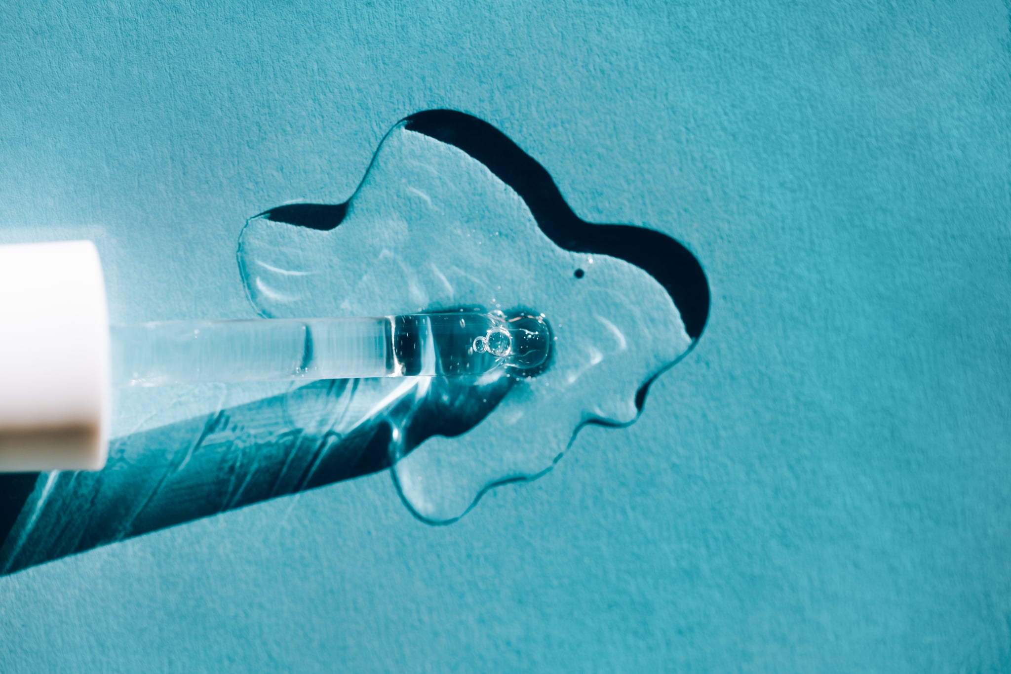 Closeup glass pipette placed in puddle of transparent lotion for spa procedure on blue background