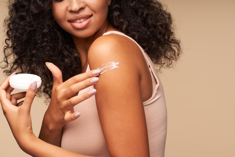Woman applying cream on shoulder while standing against brown background. Focused on lips