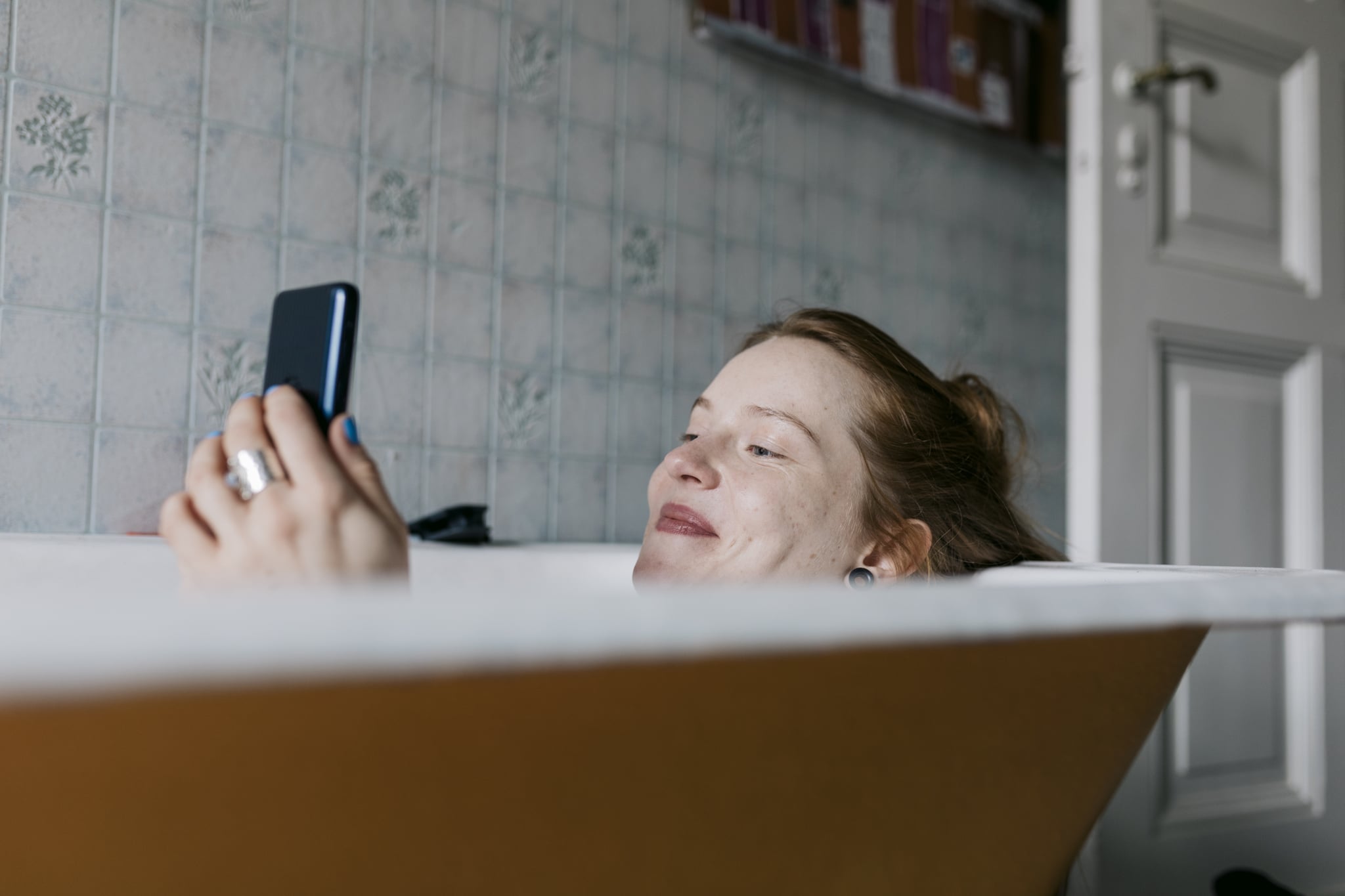 A woman taking a bath and smiling while messaging someone she met on a dating app using her smartphone.