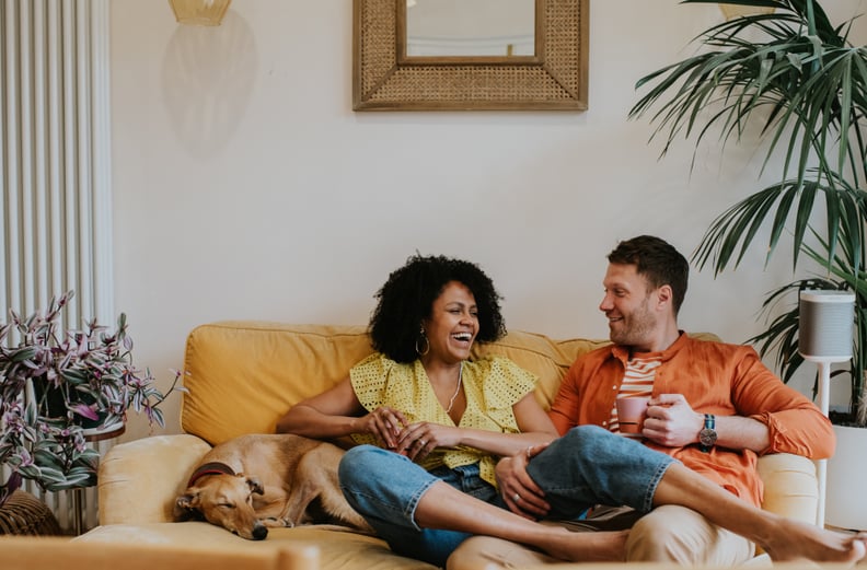 A beautiful black woman sits with her leg draped over her male partners thigh. They are comfortable and happy in a stylish living room on a soft, velvet, yellow sofa. They giggle. A lurcher dog is curled up and naps beside them. Long wall mounted modern r