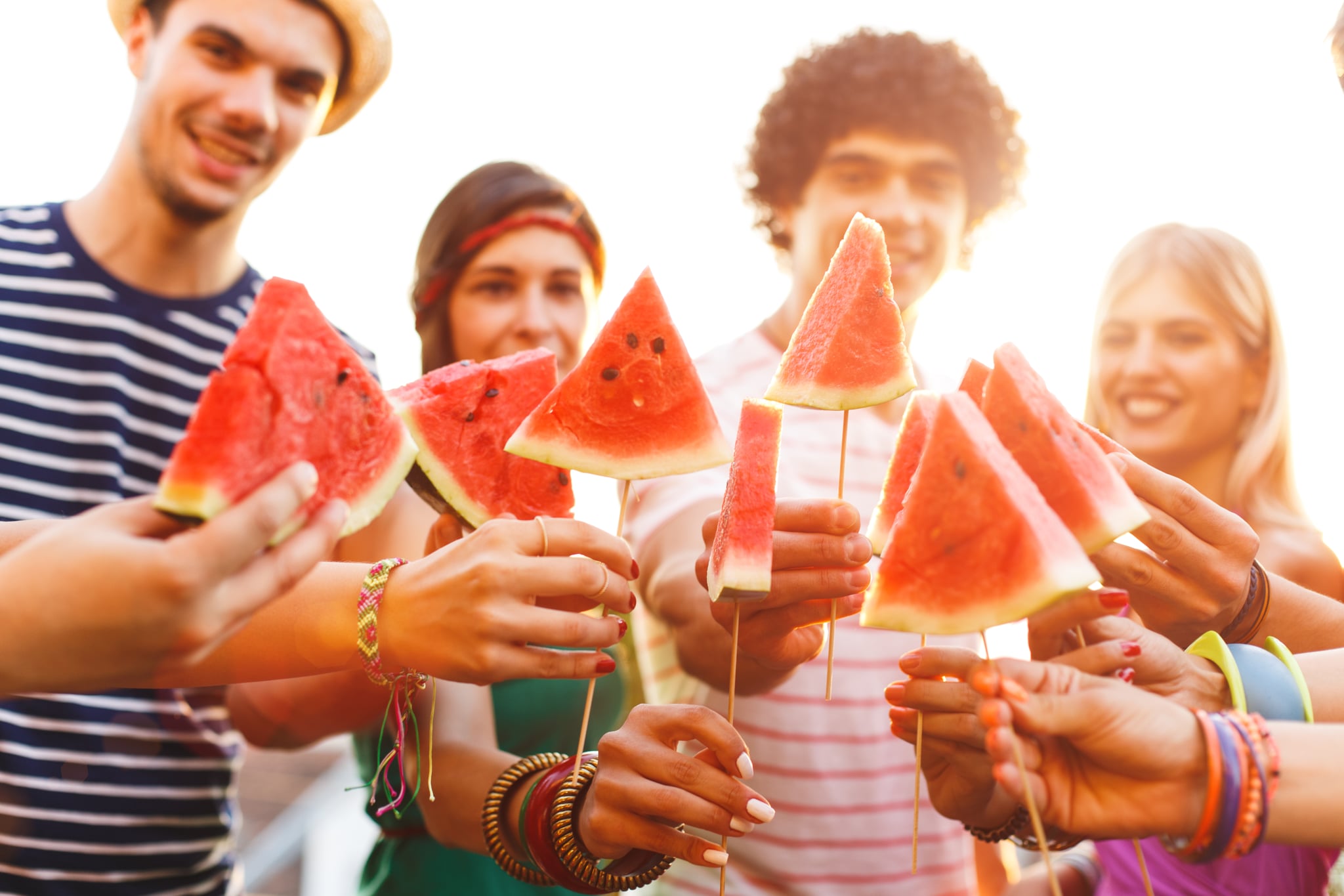 Group of young friends holding watermelon slices on a wooden stick on rooftop party, having fun, toasting.