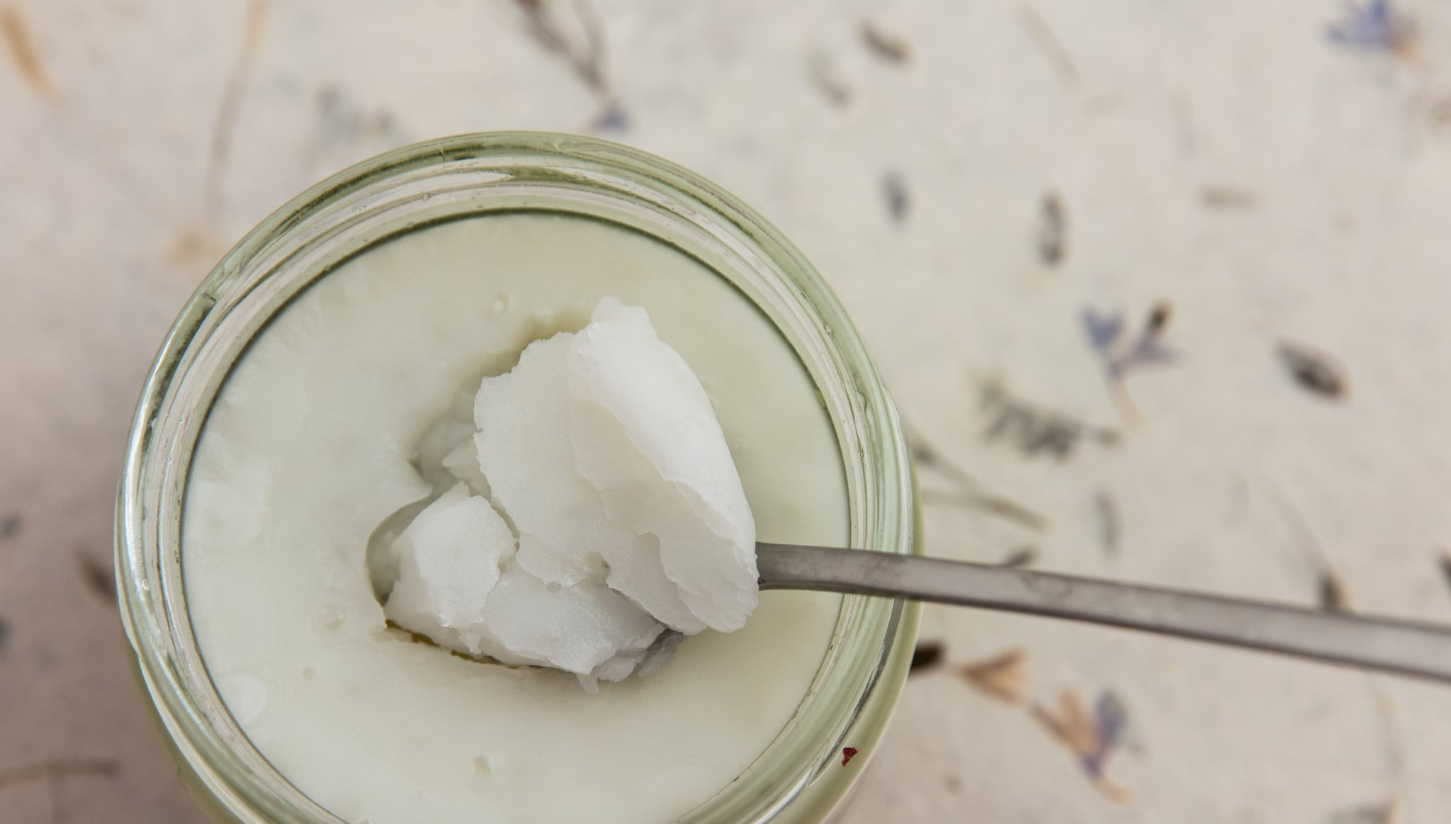 Coconut oil in a glass jar. Spoon with solidified oil. Out of focus decorative surface. High point of view. Light effect.