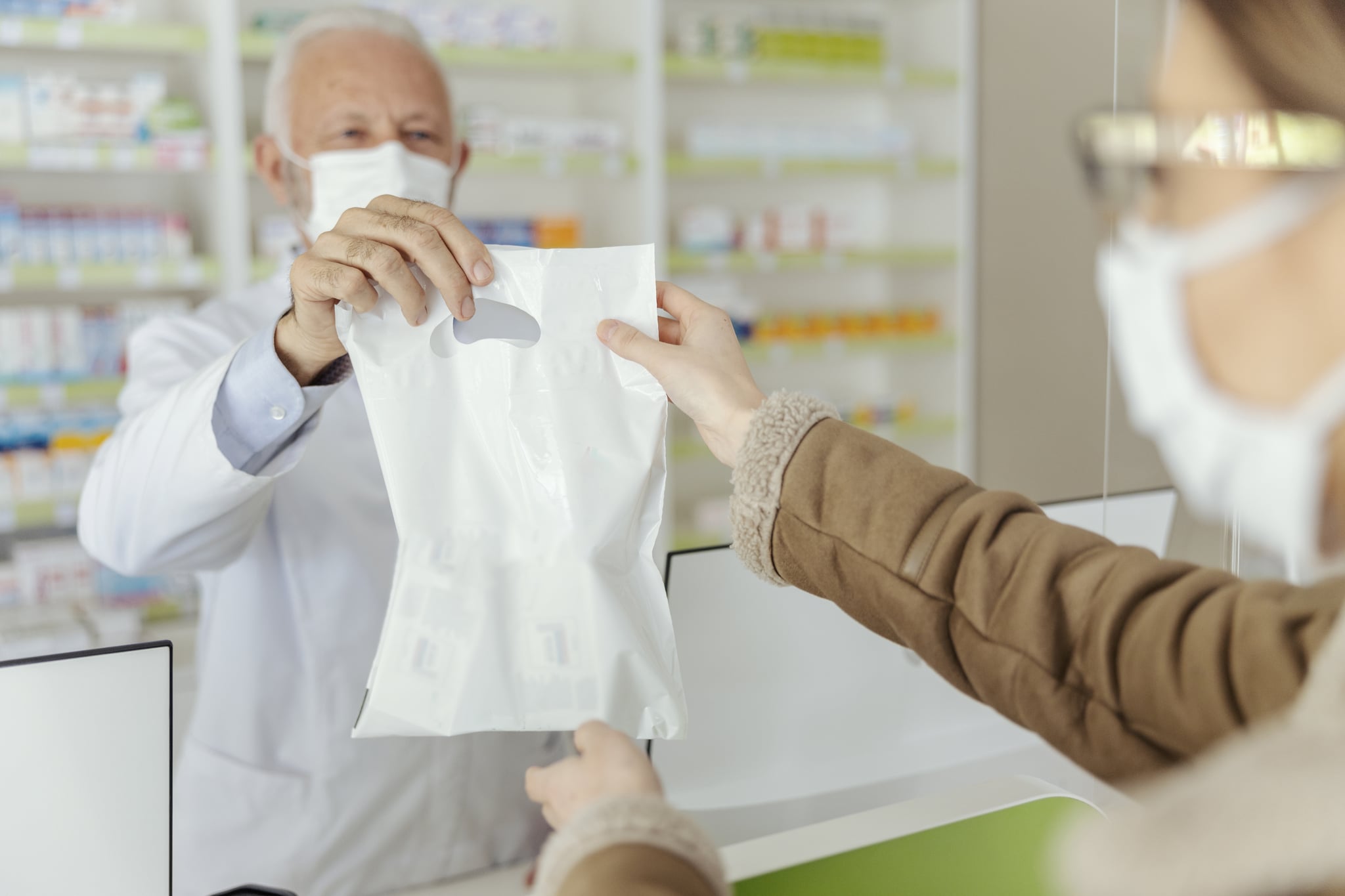 Sale of prescription drugs. An elderly male pharmacist and dressed in a white uniform with a protective mask on his face in a plastic bag of medicines and supplements delivered to the customer