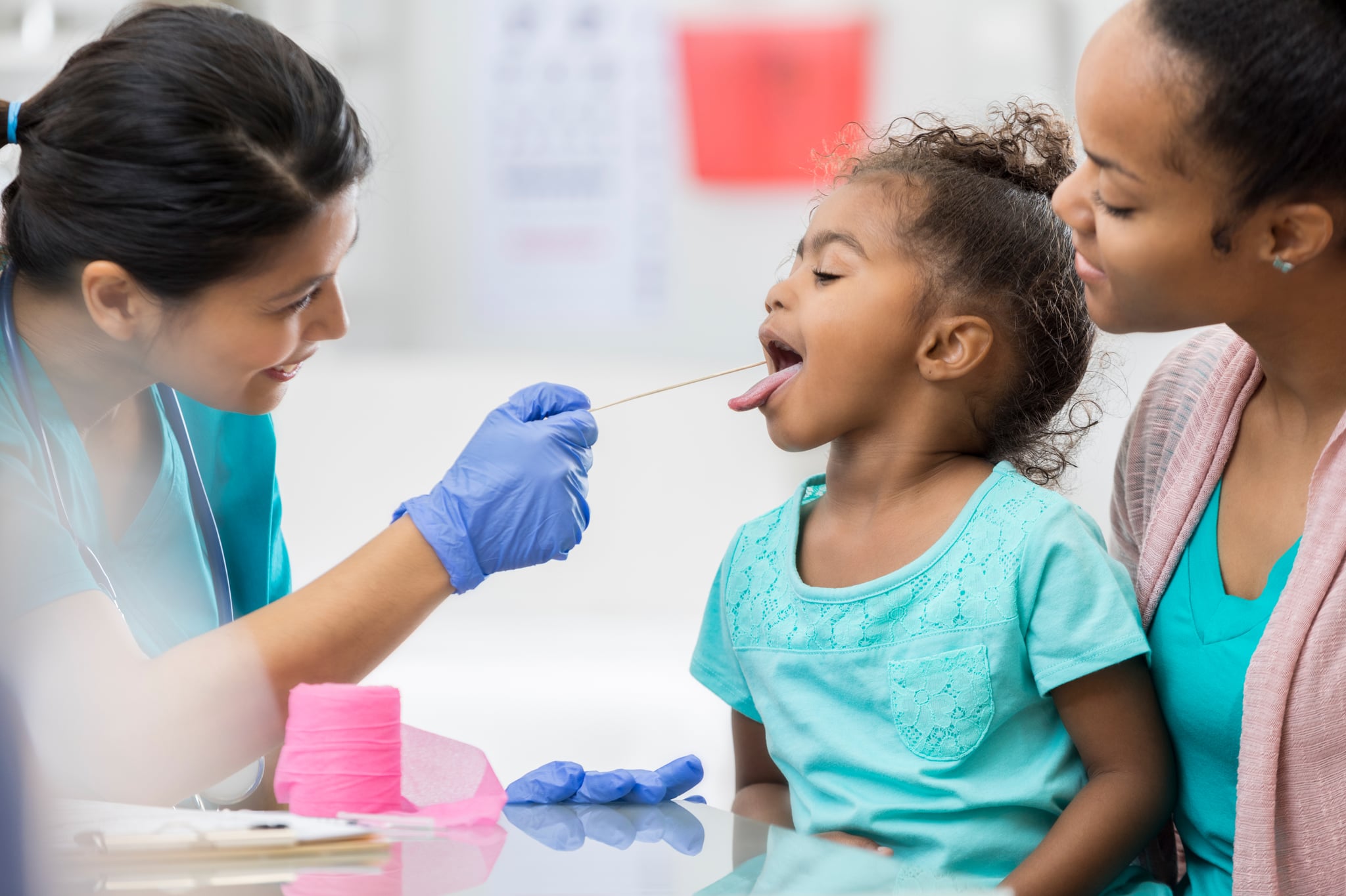 A brave little preschool age girl sits in her mother's lap at a table in the doctor's office.  She opens her mouth wide for the nurse to swab her throat to check for strep.