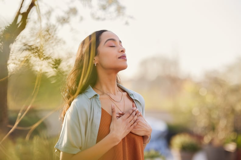 Young latin woman with hand on chest breathing in fresh air in a beautiful garden during sunset. Healthy mexican girl enjoying nature while meditating during morning exercise routine with closed eyes. Mindfulness woman enjoying morning ritual and relaxing