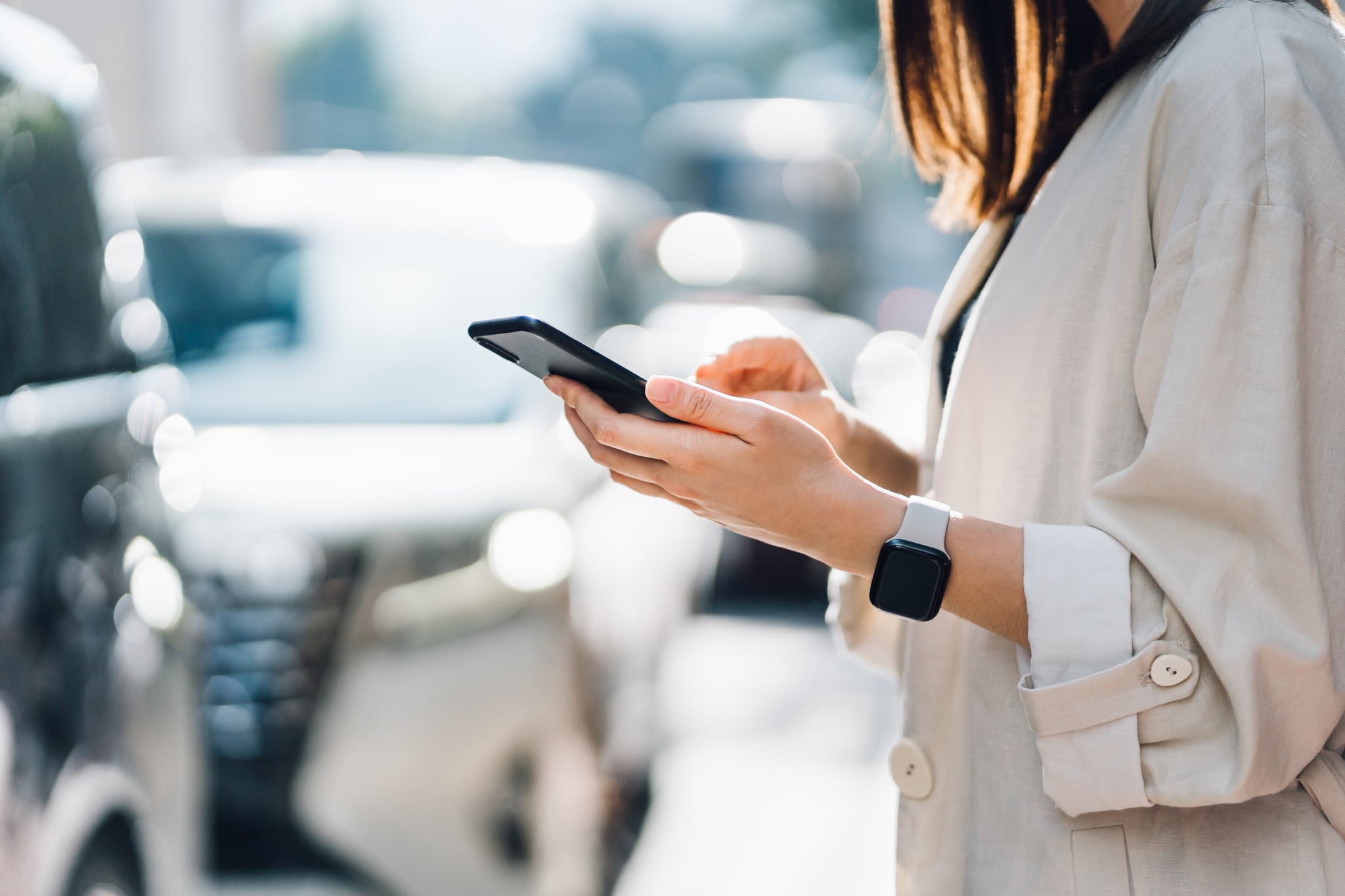 Cropped shot of chic young Asian woman using mobile app device on smartphone to order a taxi pick up service by the urban road in downtown city street. Speedy and trustworthy service. Carsharing and business on the go themes