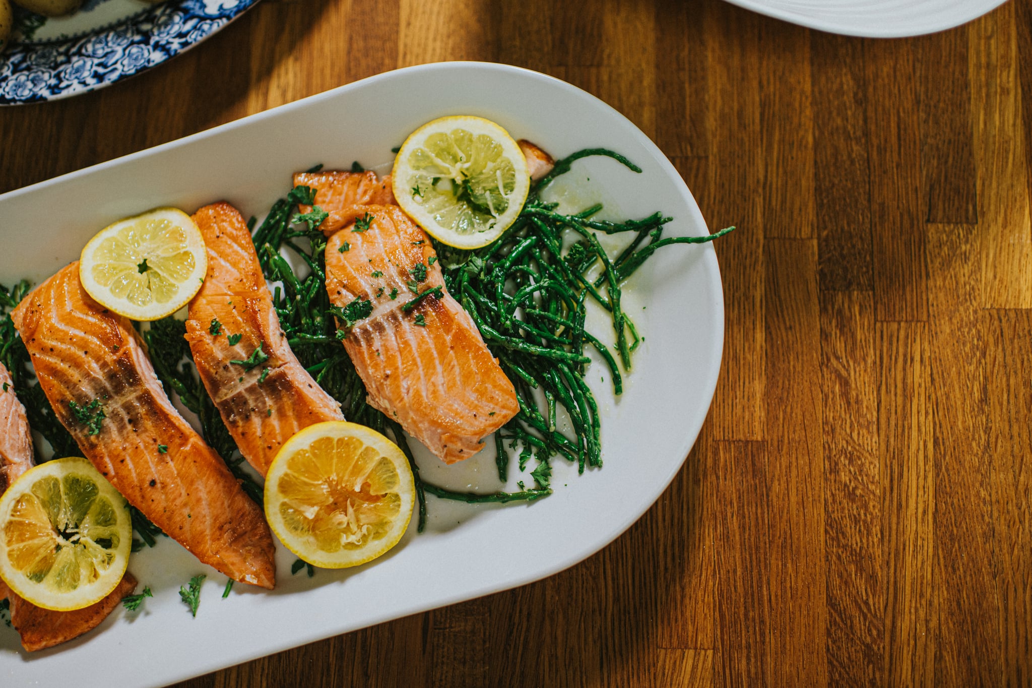 Top down image of four salmon darns on a white long oval serving ceramic plate on a wooden table. It rests on a bed of samphire. Lemon slices garnish the fish.
