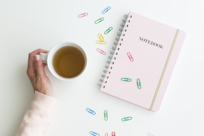 Woman holding a mug of hot fresh tea with her hand alongside a pink notebook with scattered colorful paperclips over white viewed from overhead