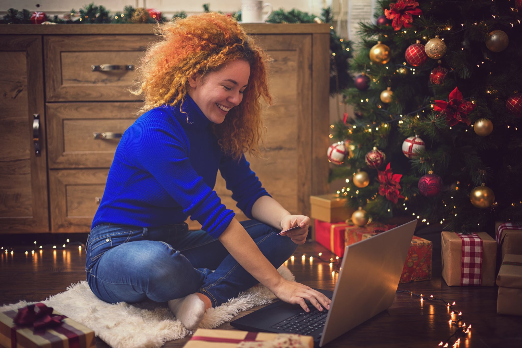 Young beautiful girl with red curly hair ,enjoying at home in a cozy Christmas atmosphere while using lap top for online shopping.