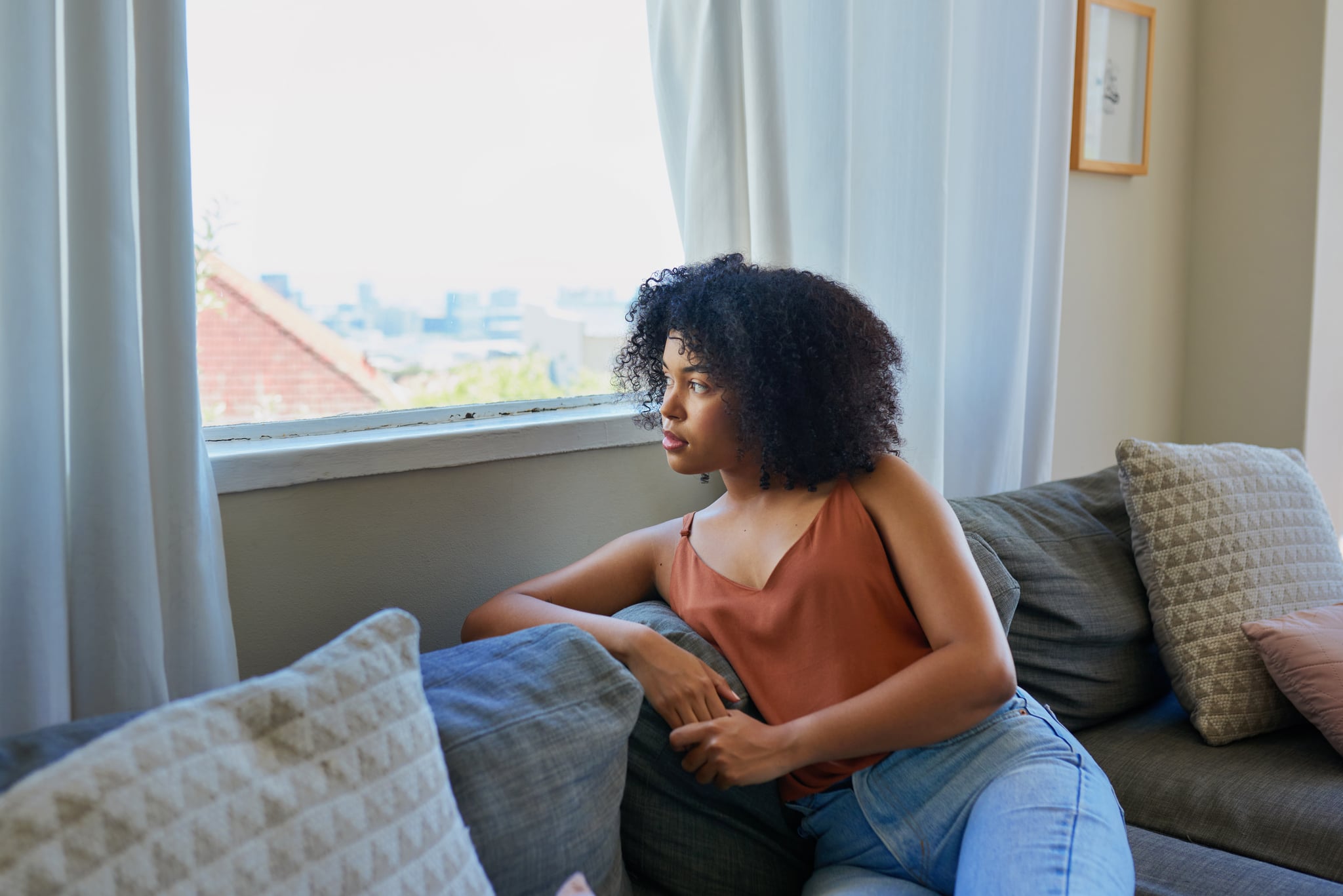 Shot of a young woman sitting on the couch, looking out the window, and dissociating.