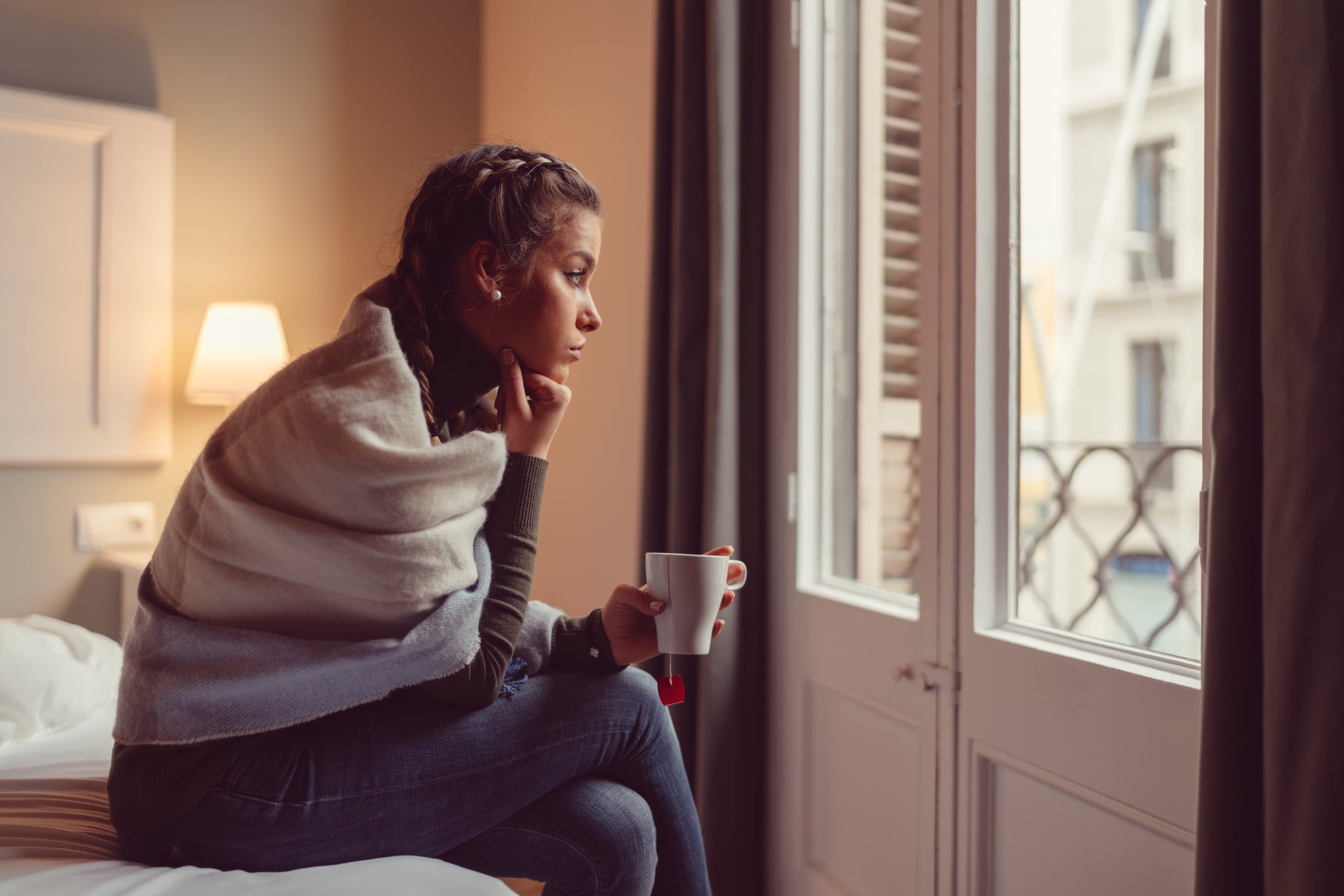 Young woman sitting at the bed with cup of tea and looking through the window