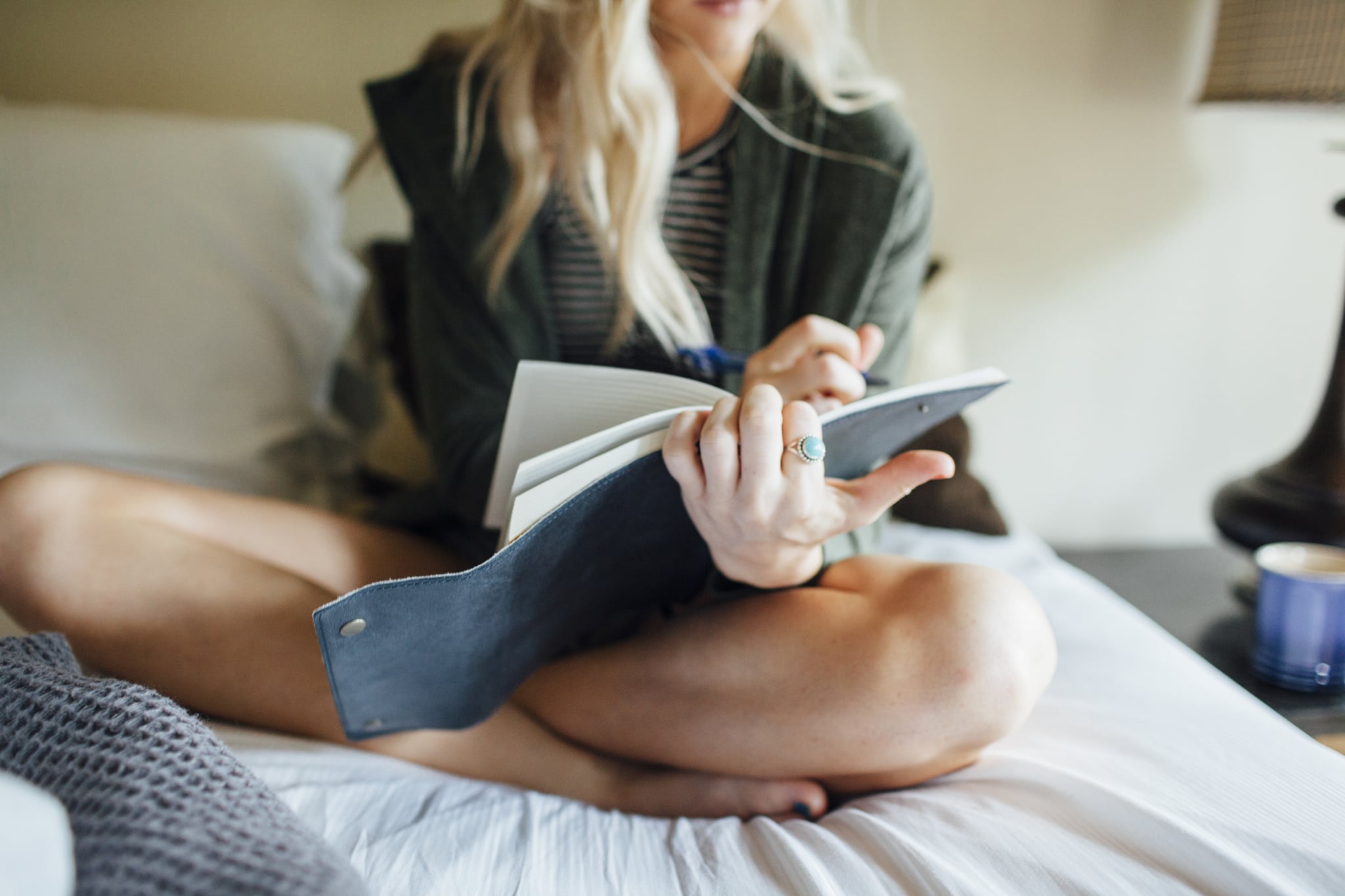Woman with blonde hair sitting with her legs crossed on a bed while writing in a blue notebook.