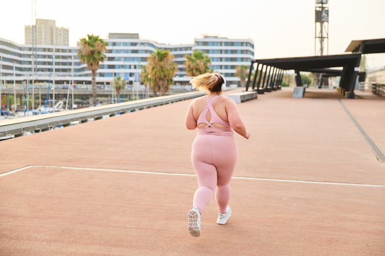 Full body back view of young plus size female listening to music on headphones while jogging in city during weight loss training. Working to take care of your body and health