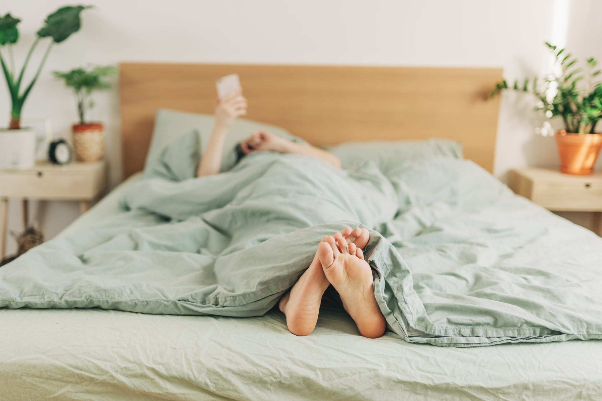 A woman alone in her bed looks at her phone while her bare feet stick out of blue sheets.