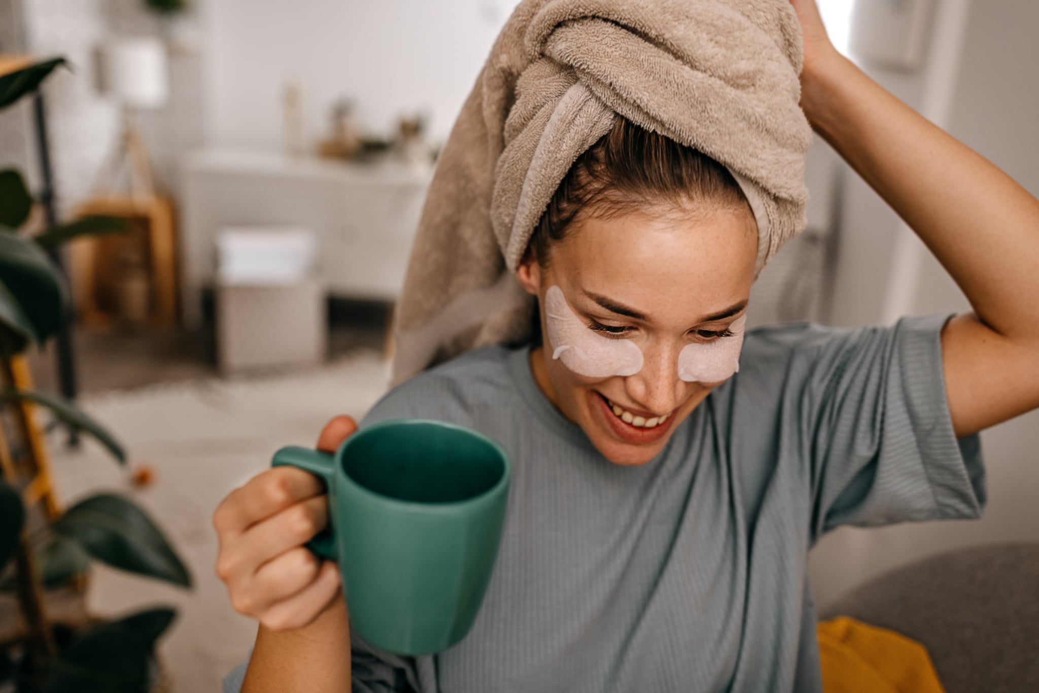 Pretty young woman with healthy skin applying cosmetic patches under eyes drinking coffee after shower with towel on hair looking away