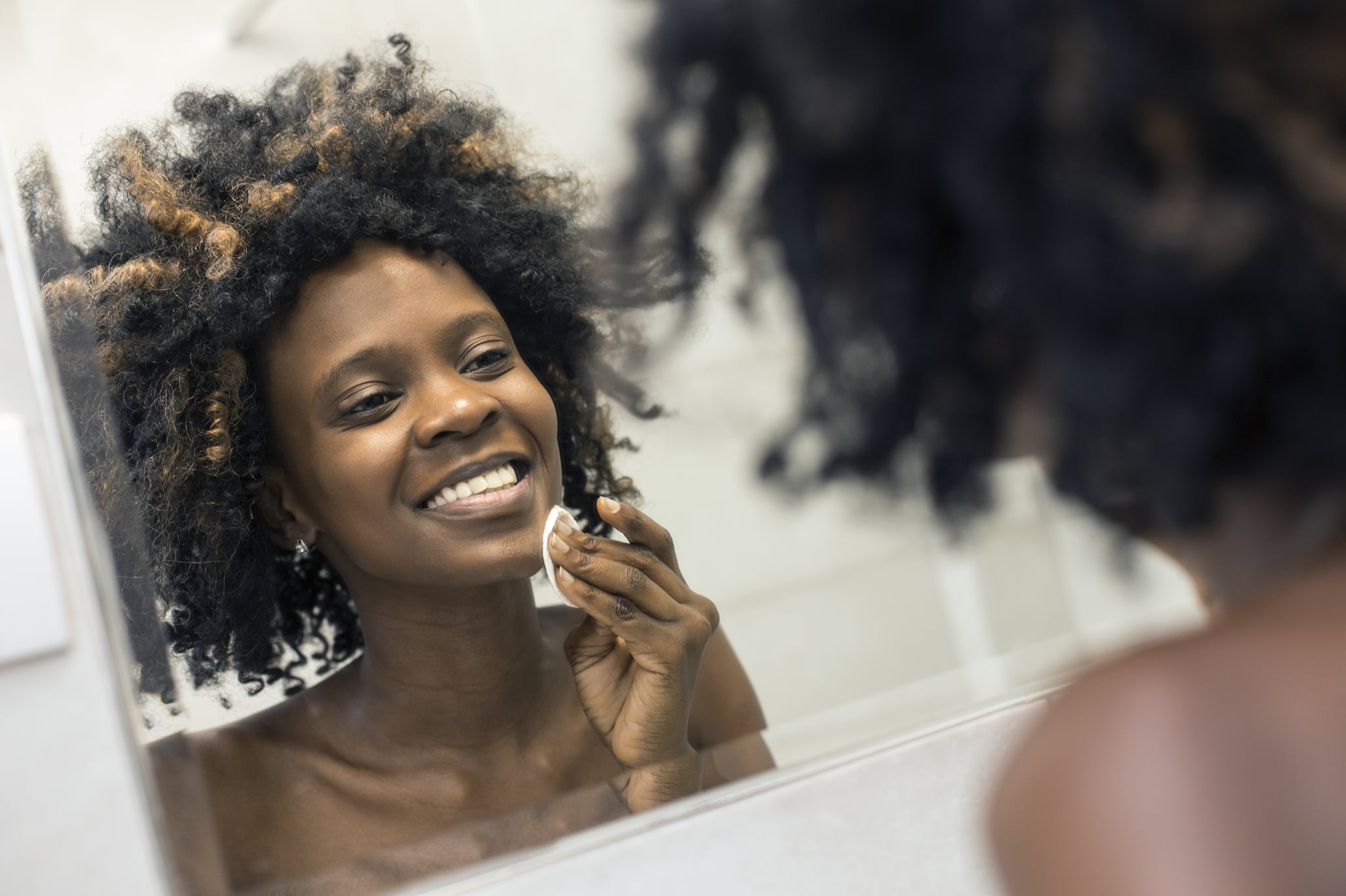 Young woman cleaning her skin in bathroom mirror with a cotton pad. About 25 years old, African female with curly hair.