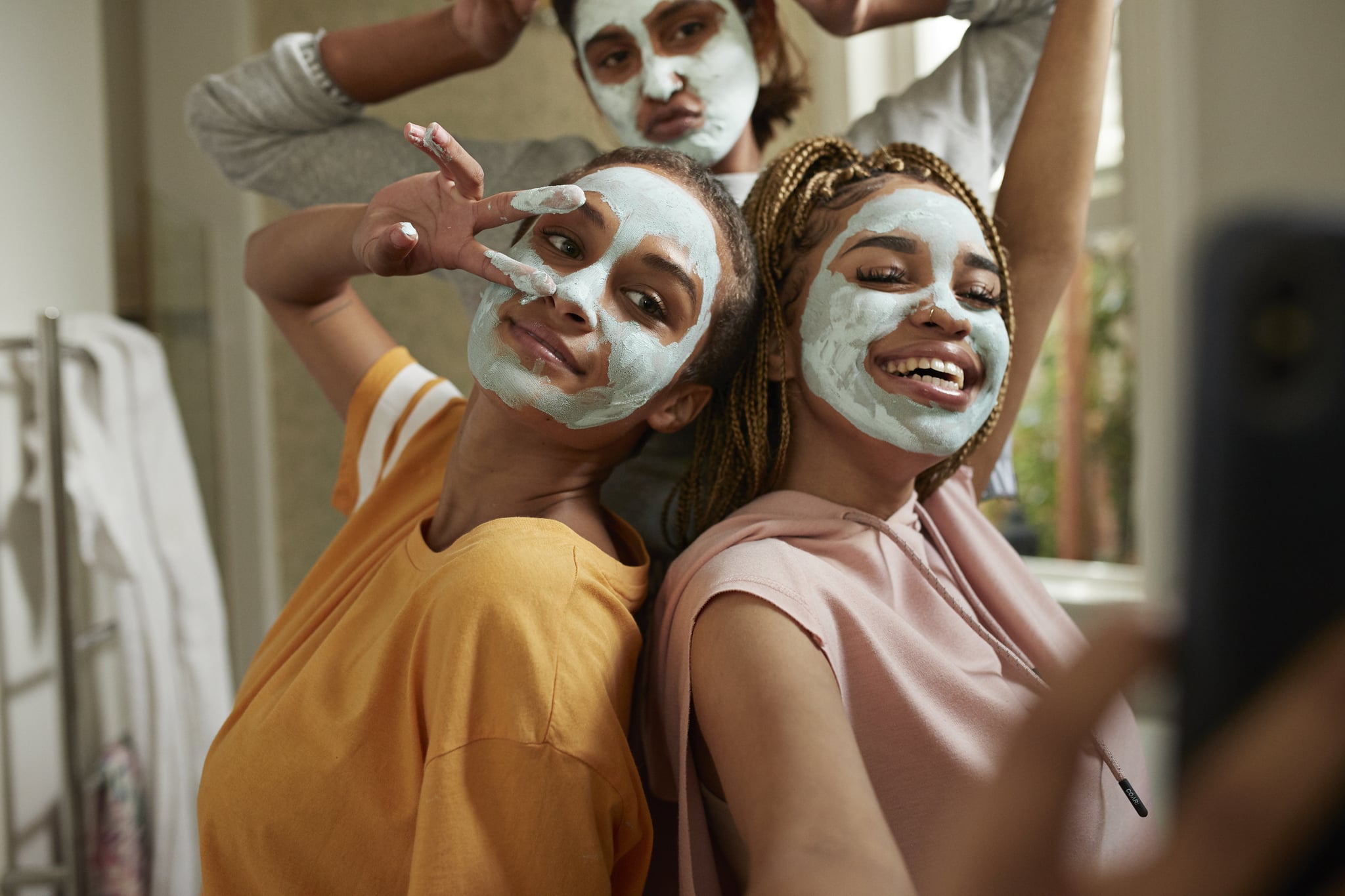 Happy young woman taking selfie with female friends wearing facial cream in bathroom at home