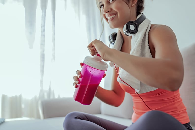 关闭年轻健康女性的形象与耳机喝蛋白奶昔后,坐在客厅的锻炼。年轻的女运动员运动服装坐着,喝蛋白质鸡尾酒摇