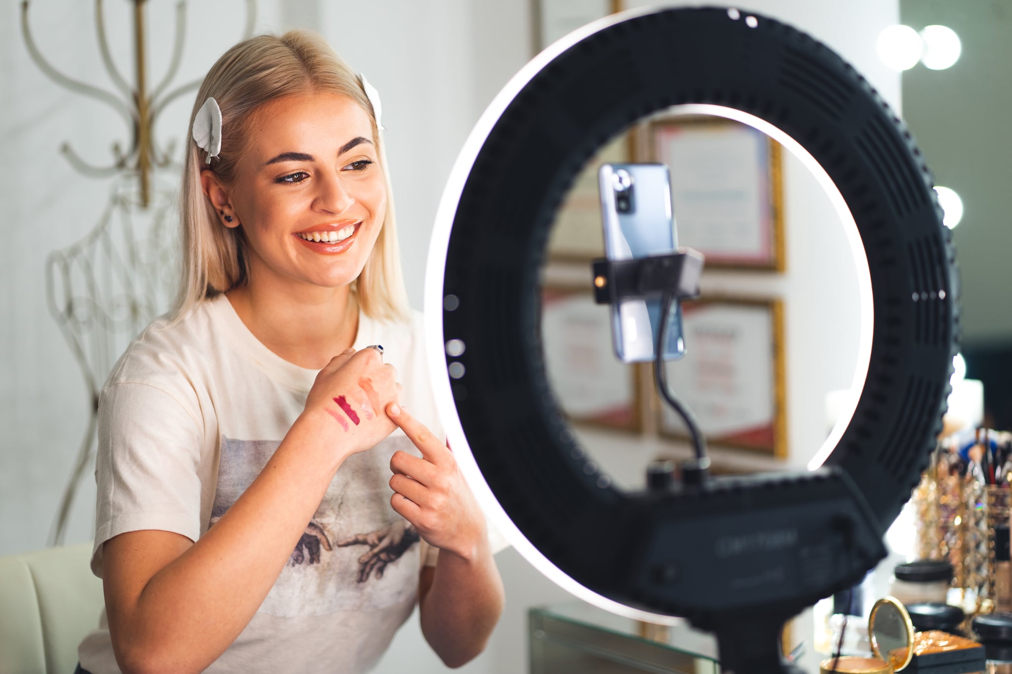 A young female beauty blogger is recording a vlog video on a smartphone using the ring light. Shot behind the ring.