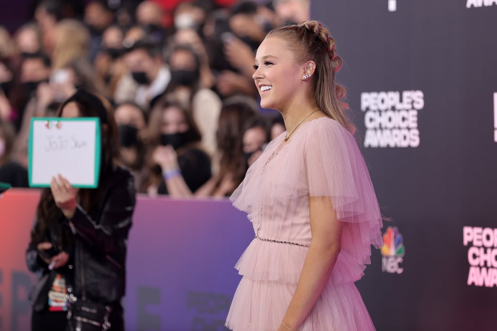 JoJo Siwa continues to shape her style. The 18-year-old YouTube personality and recent Dancing With the Stars competitor has been swapping her neon separates for elegant ballgowns, and her appearance at the People's Choice Awards on Dec. 7 follows suit. 
JoJo attended the award show wearing a pink midi-length dress by Jenny Packham with tulle layers and a beaded neckline. (With its pleated sleeves, in fact, the dress actually brought to mind Glinda the Good Witch from The Wizard of Oz!) JoJo completed the sweet look with sparkly Christian Louboutin pumps, a gold necklace, and a colourful earring stack. In support of her fans, JoJo also wore a J Nation necklace wrapped around her wrist as a bracelet. 
This appearance comes shortly after JoJo attended the American Music Awards in a glamorous black gown. "I've kind of had this transition while being on DWTS," she said on the Nov. 21 award show carpet. "I've gone from dressing like how I normally dress to looking more adult, and I was like, 'I want to branch out of my comfort zone.'"