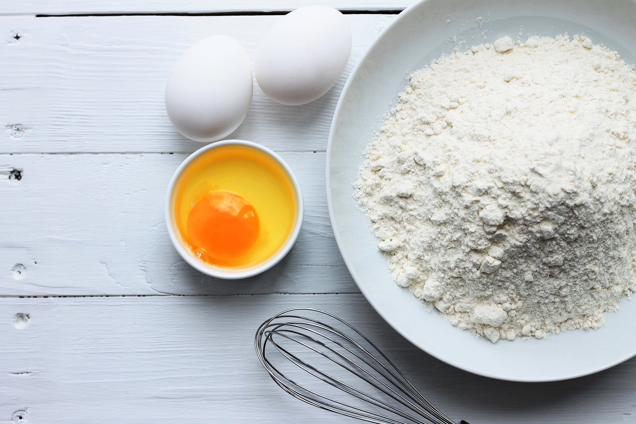 A plate with flour, eggs, a whisk on a white wooden background. Pancakes ingredients. Egg in the bowl. First step of cooking.
