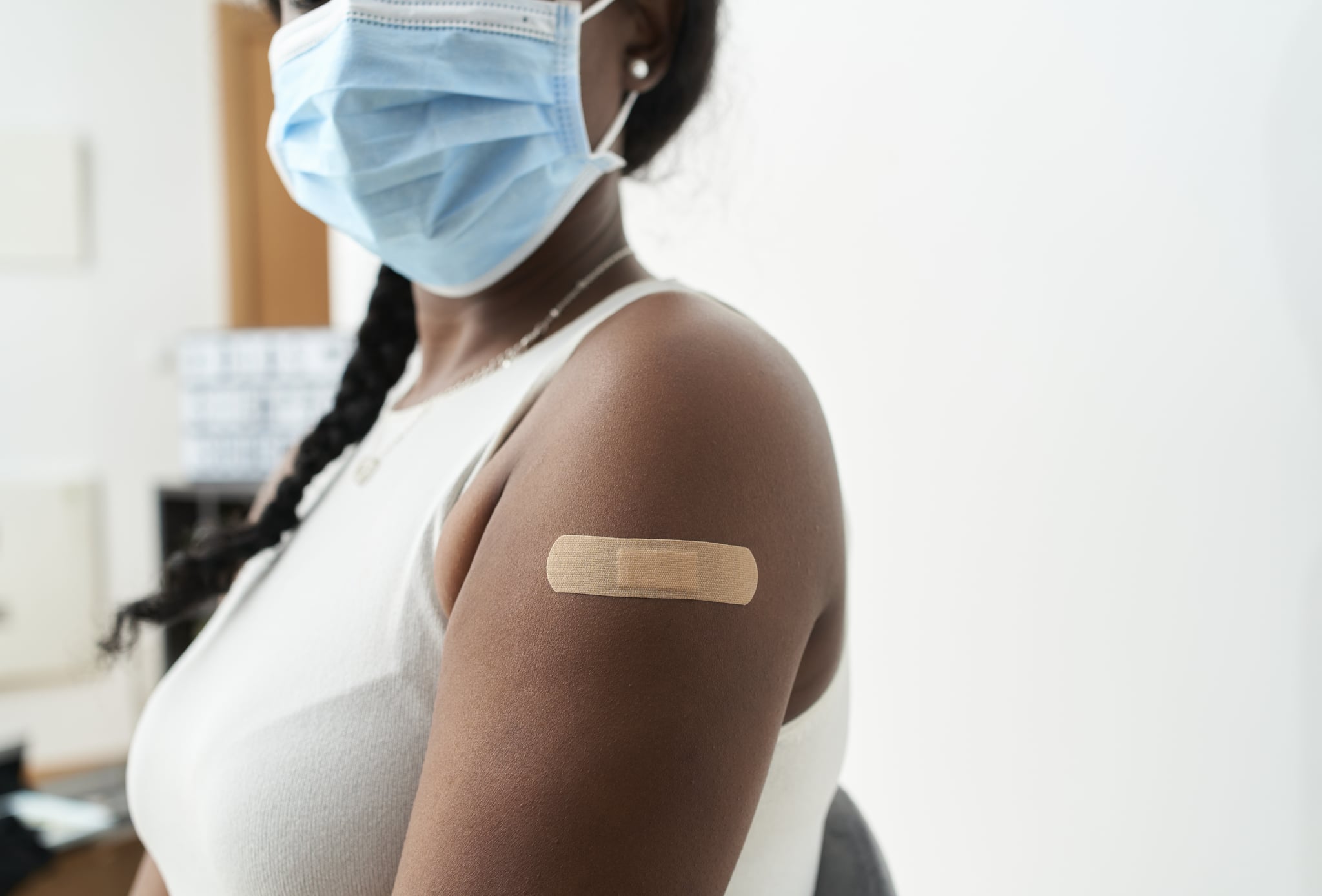 Black woman with braid in her hair with white shirt and mask showing the band-aid on her arm after vaccination, with white background. Concept of covid