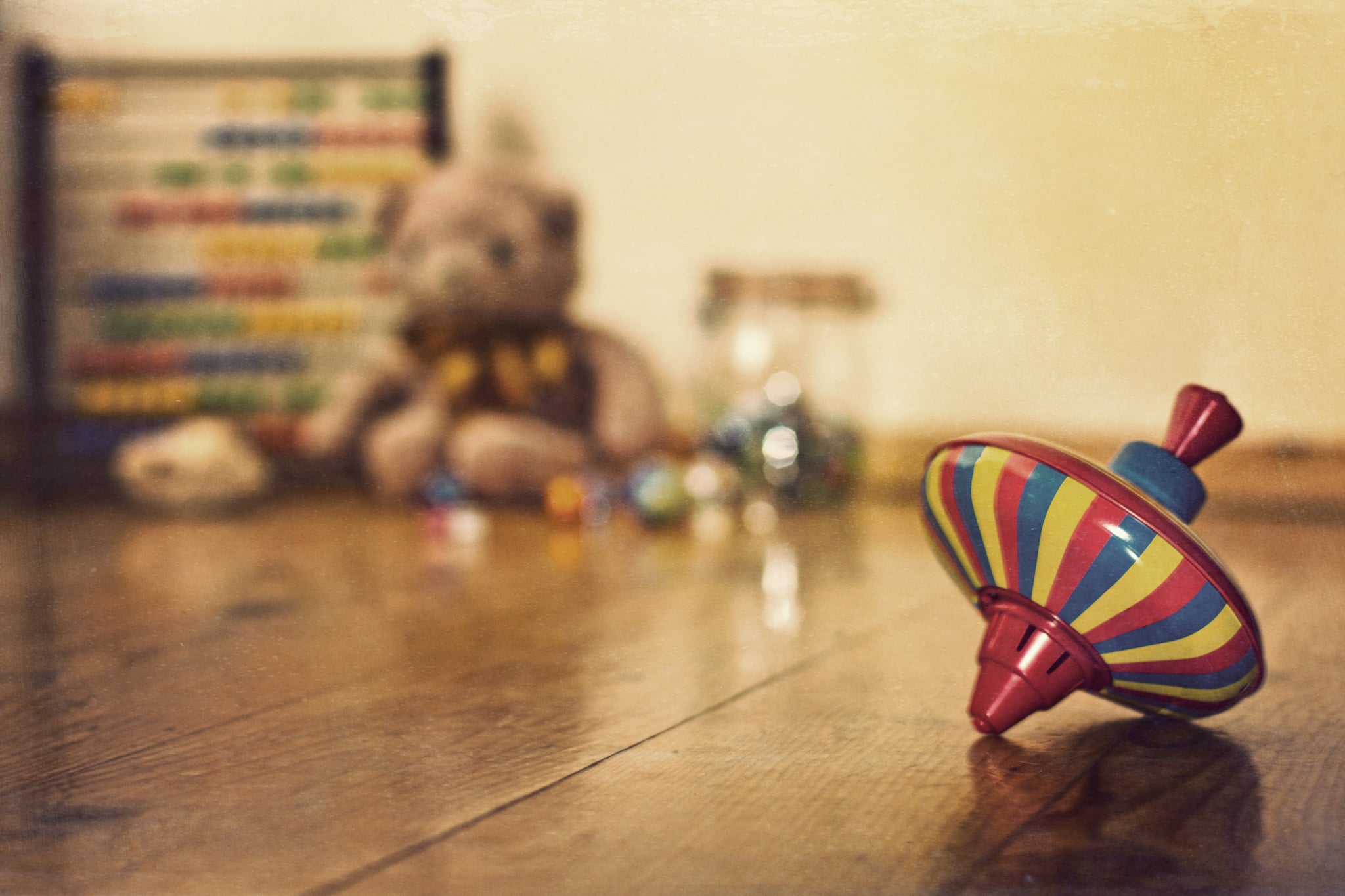 An old fashioned tin spinning top in the foreground with a collection of old fashioned toys in the background including an abacus, teddy bear and marbles.