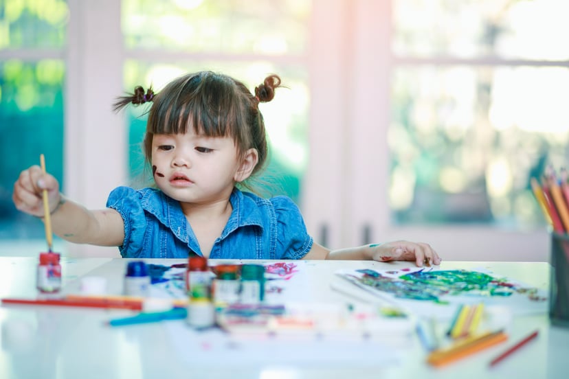 little girl painting with paintbrush and colorful paints on desk background