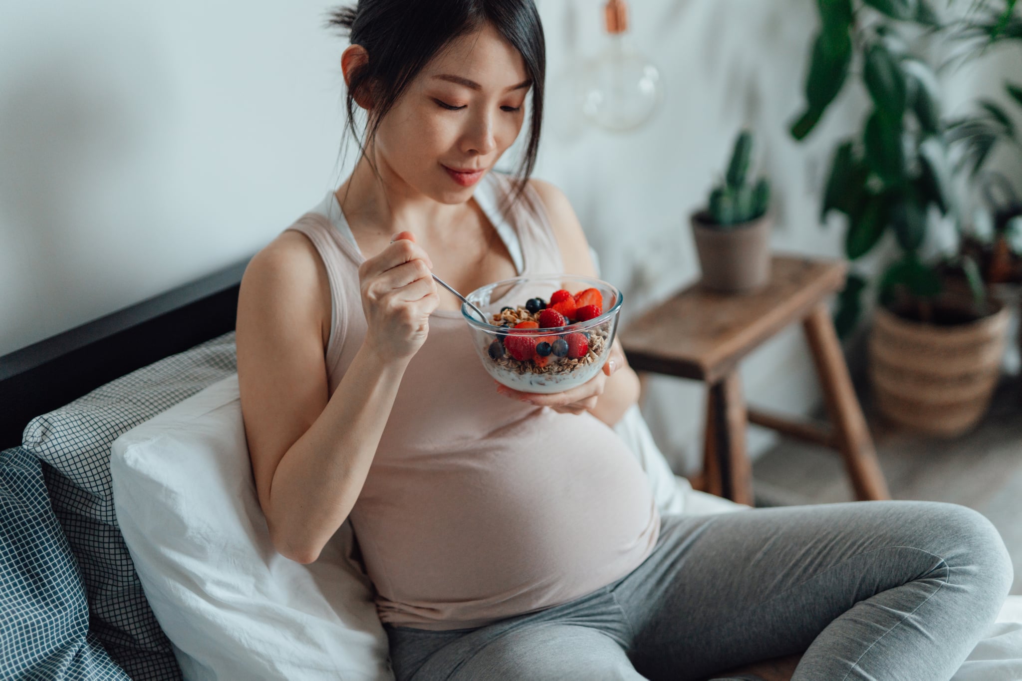 亚洲孕妇吃一碗麦片和酸奶混合浆果,坐在床上。怀孕期间吃好。健康的生活方式在怀孕。