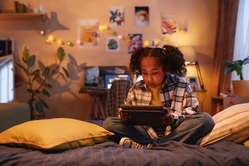 A young girl with pigtails uses a digital tablet while sitting on a bed in cozy warm light.