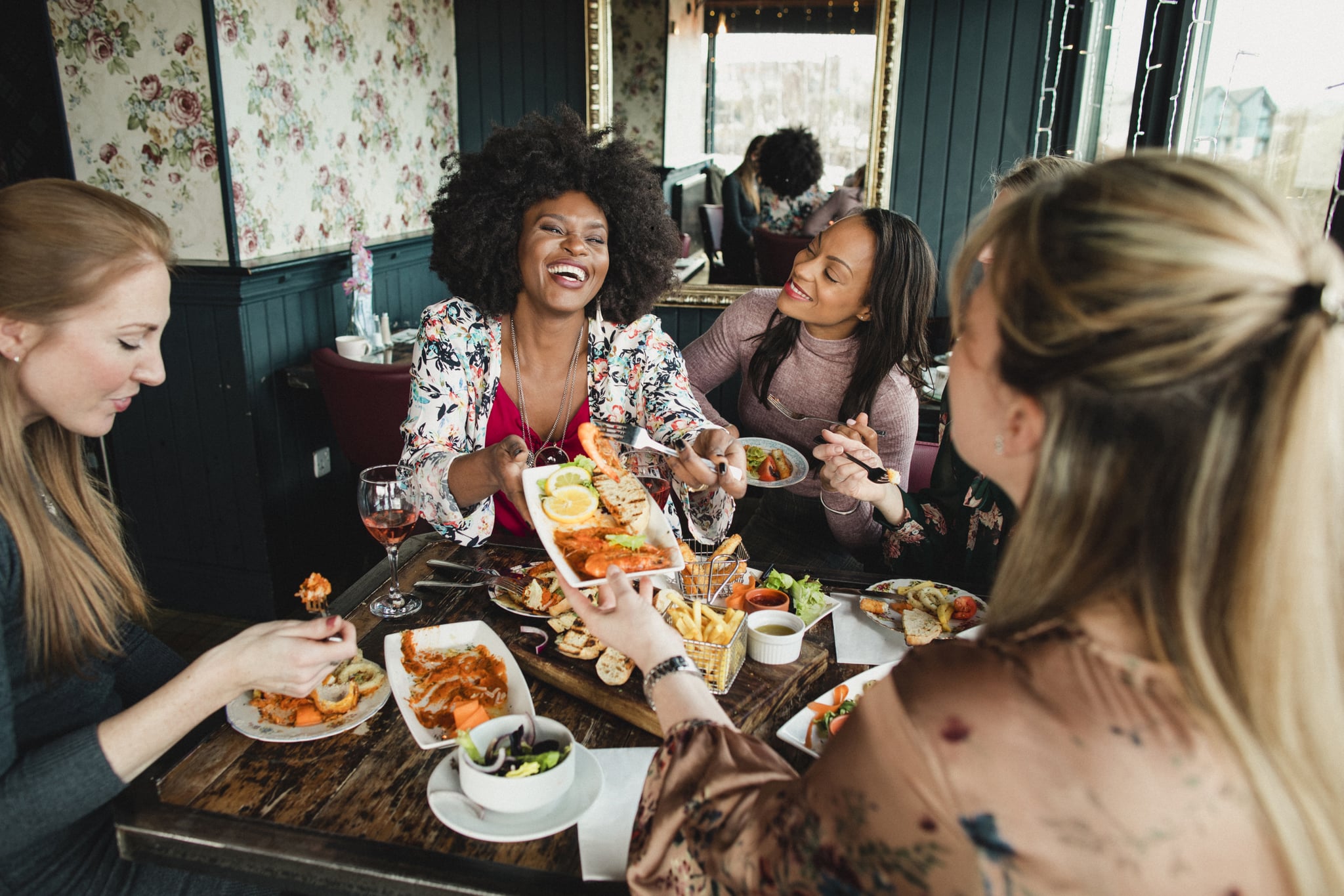 A group of women are enjoying a meal in a restaurant with rose wine. They have a king prawns sharing platter , one woman can be seen passing her friend a plate of food. Mixed ethnic group
