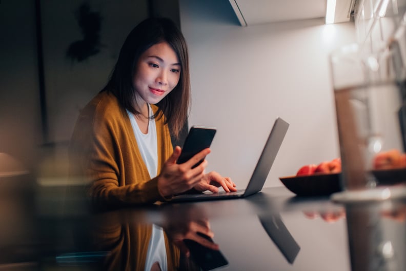 Beautiful young Asian woman looking at smartphone while working with laptop in the kitchen at home in the evening.