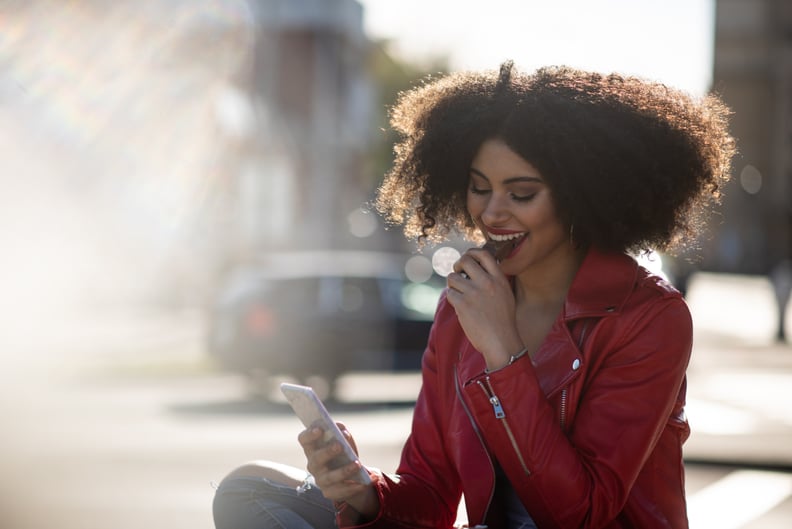 Smiling teen black curly hair girl looking to phone and eating chocolate snack in sunny street