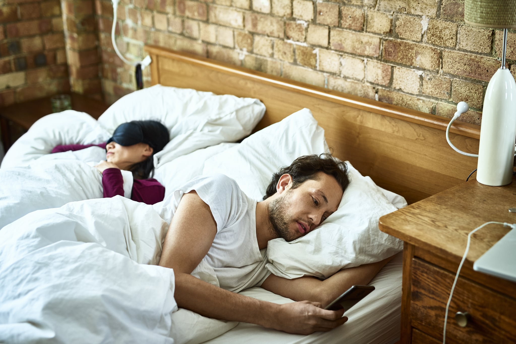 Mature man sending text message as his girlfriend sleeps in the bed, serious expression on face