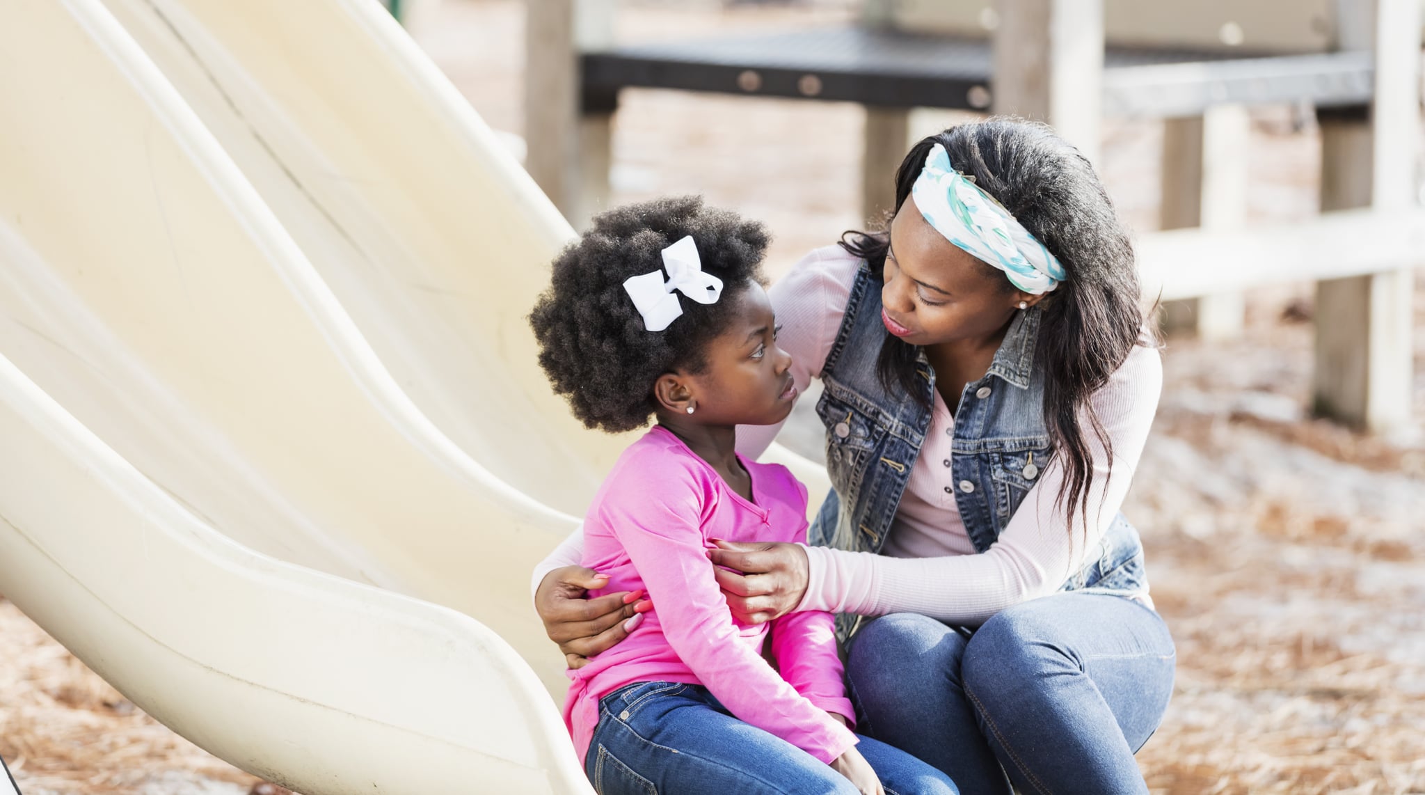 A young African-American woman helping her 5 year old daughter who is sitting on a slide on a playground with a sad expression on her face.
