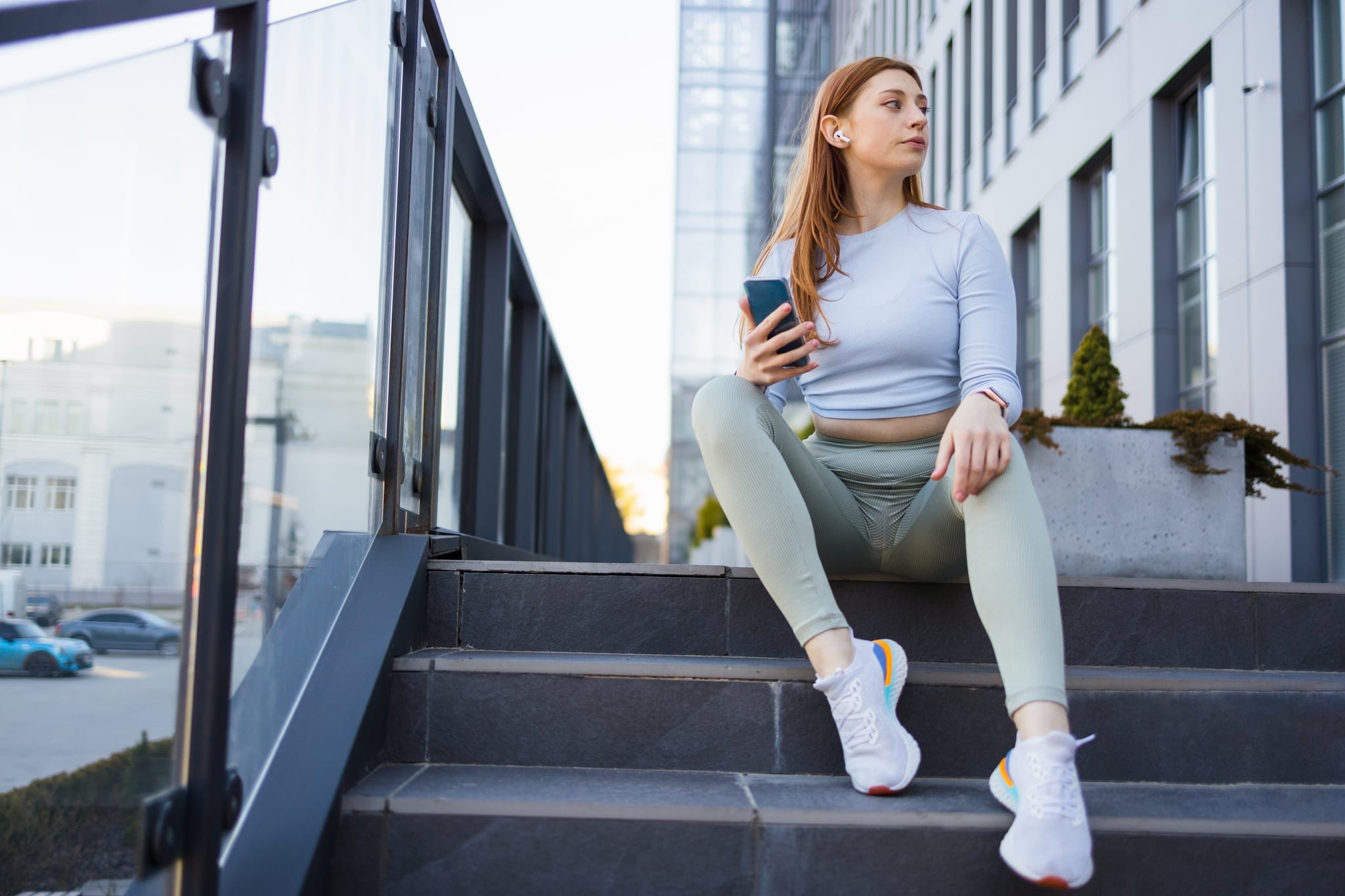 Smiling sporty girl with earphones using phone while sitting down on floor after hard workout