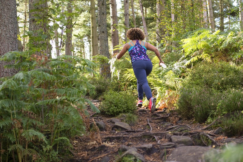 Rear view shot of a woman running through a forest across extreme terrain. She is wearing sportswear and is carrying a waterbottle.