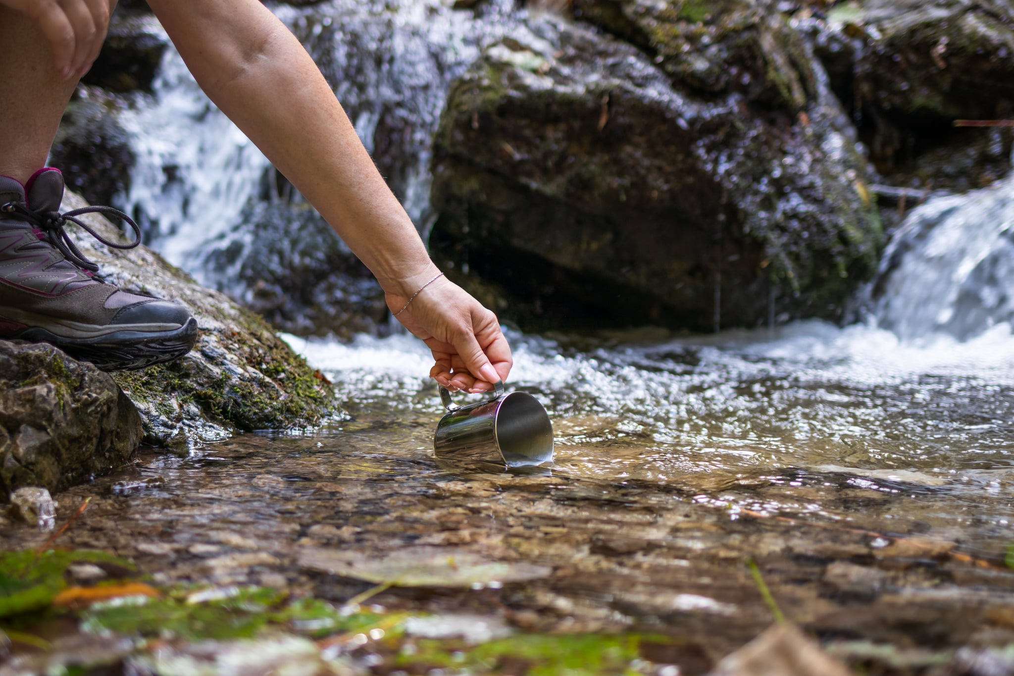 Thirsty hiker scoops fresh water into cup from stream in mountains. 