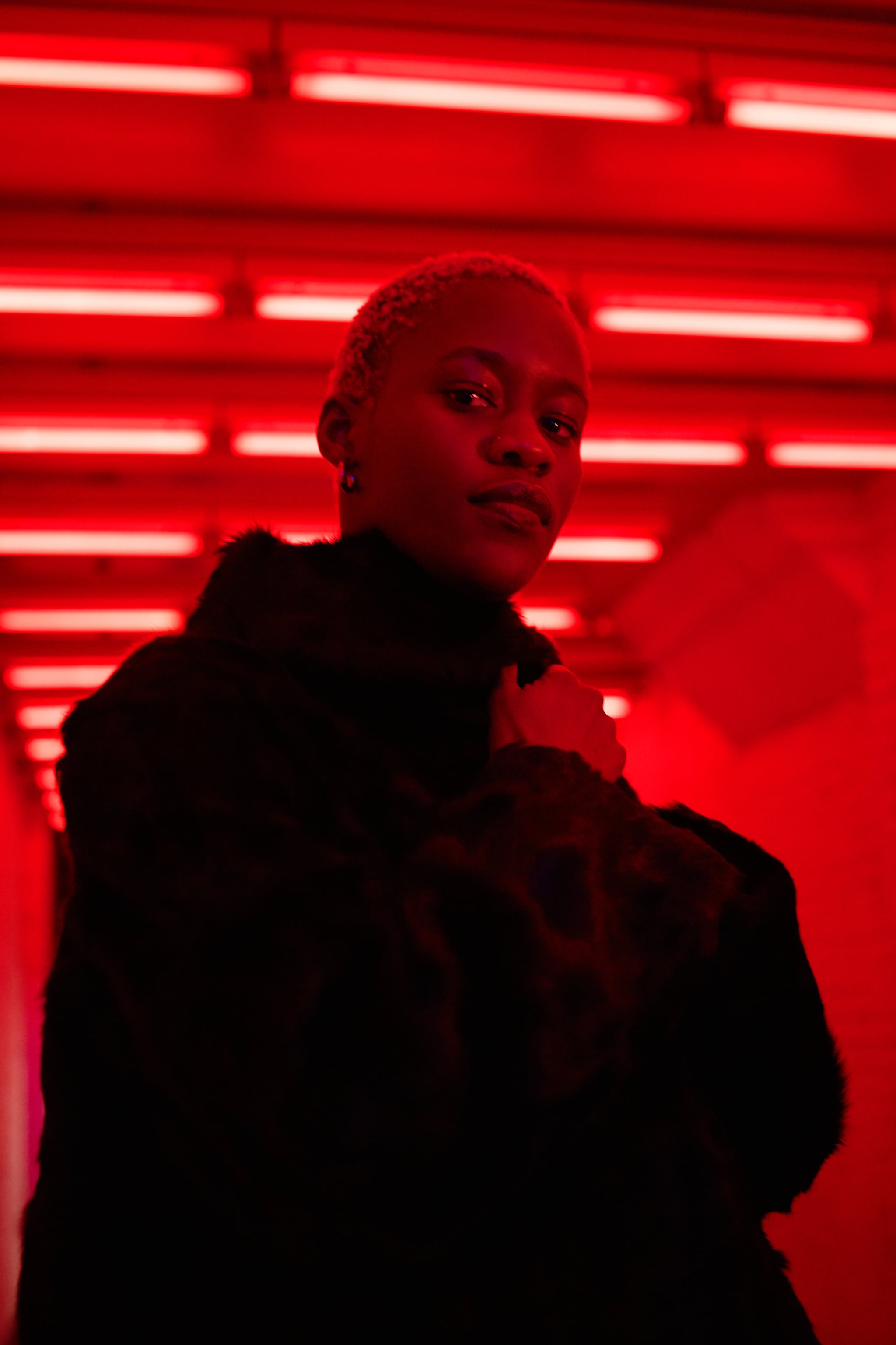 Portrait of young woman with blond short hair wearing black fur and white sweater infront of neon lights.